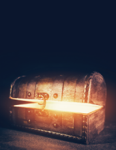 A wooden treasure box that is slightly ajar - light glows from within the box. Background is black.