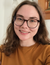 Laura Moberg smiles at camera. She has dark rimmed glasses, pale skin, and wavy brown hair that falls just below her shoulders.