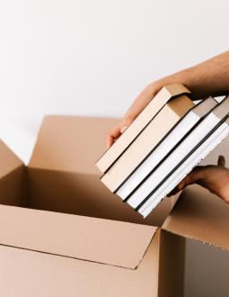 Close-up of hands packing books into a cardboard box on a white background.