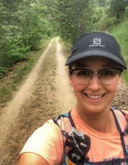 Kyle smiles into the camera. She has a black cap on her head and is wearing a tshirt and backpack. Behind her is a view of a trail.
