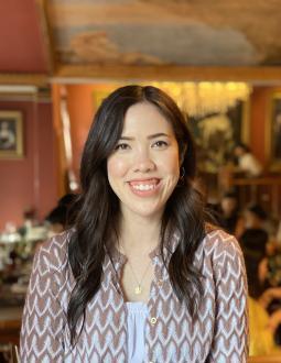 Sasha smiles at the camera, with a blurred background of a restaurant. Sasha has long black wavy hair that falls several inches below her shoulders.